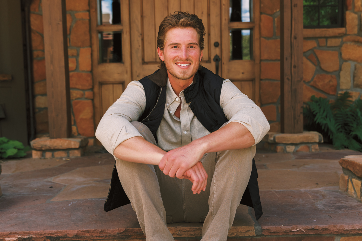 Man sitting on stone steps outside a rustic wooden doorway, wearing a neutral button-down shirt, black quilted vest, and taupe pants.