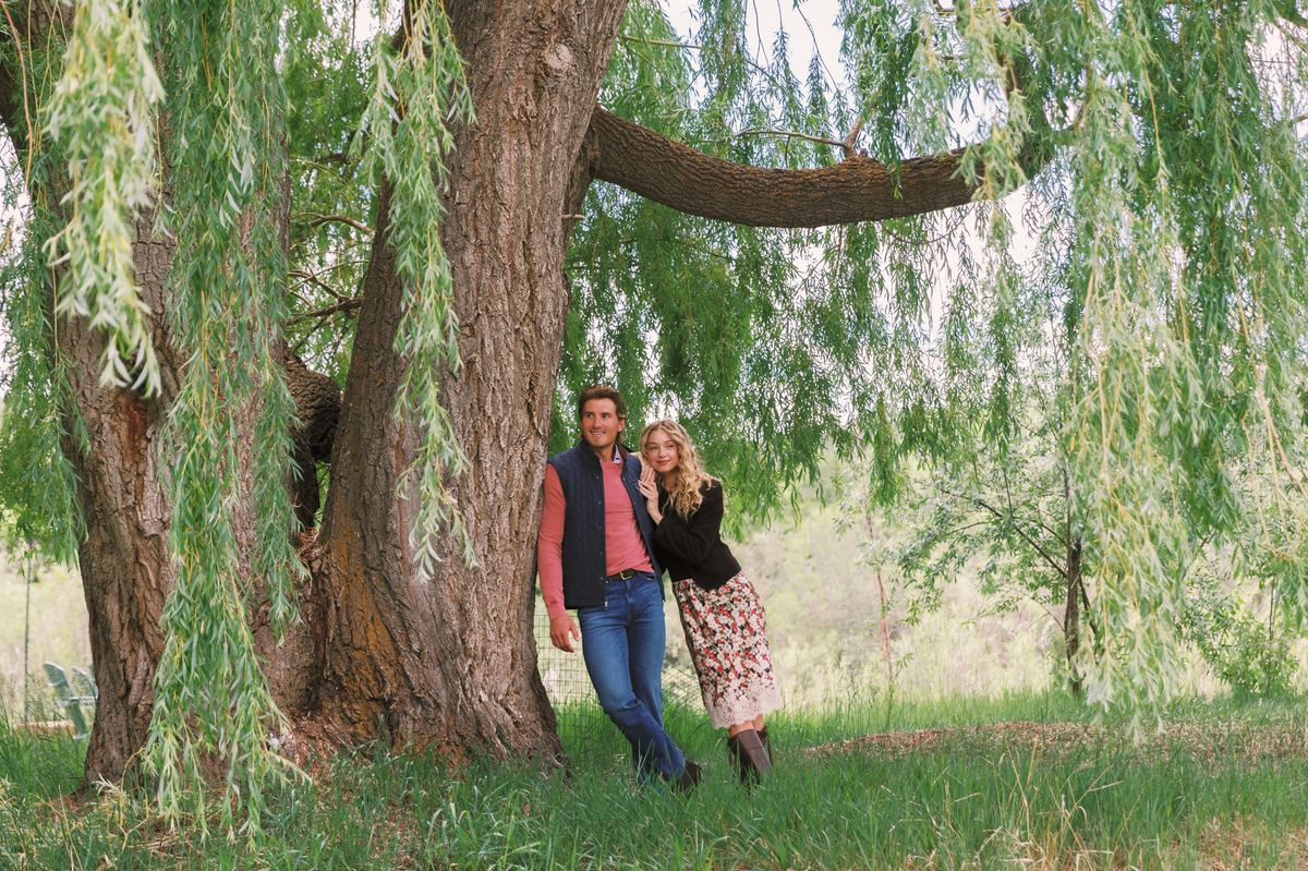 A couple standing together beneath a large willow tree in a grassy field, surrounded by lush green foliage. The man wears a red shirt, navy vest, and jeans, while the woman wears a black jacket, floral skirt, and brown boots.