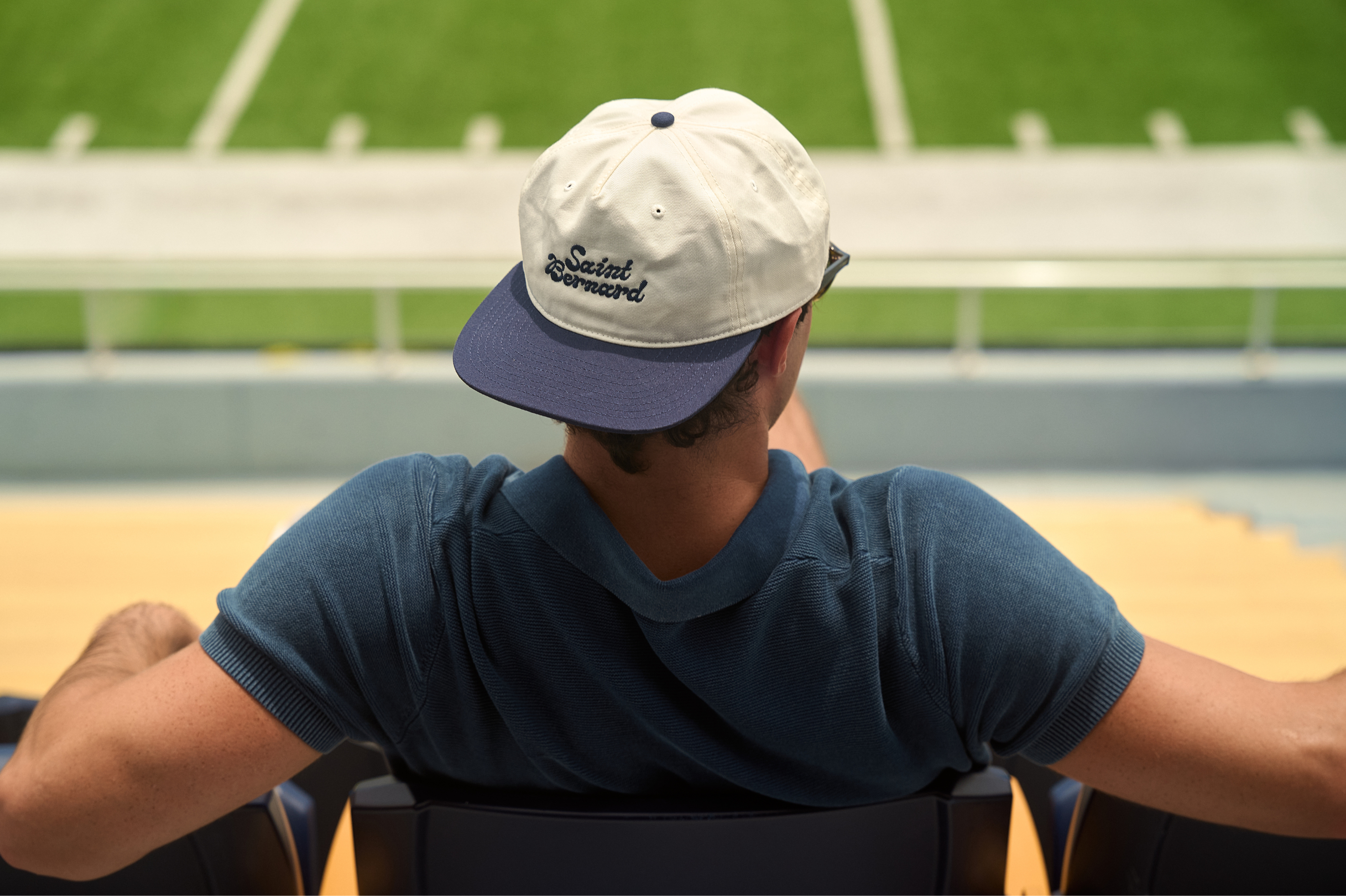 A man viewed from behind wearing a navy blue and off-white baseball cap with 'Saint Bernard' embroidered on the front, seated in a stadium.