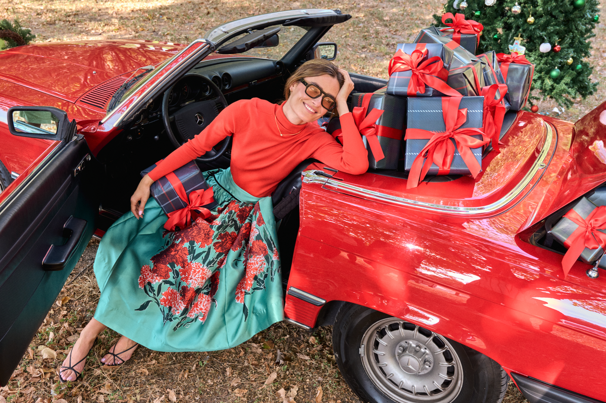 Woman wearing a red turtleneck and turquoise floral skirt sits in a vintage red Mercedes convertible surrounded by plaid-wrapped Christmas gifts with red bows, beside a decorated Christmas tree.