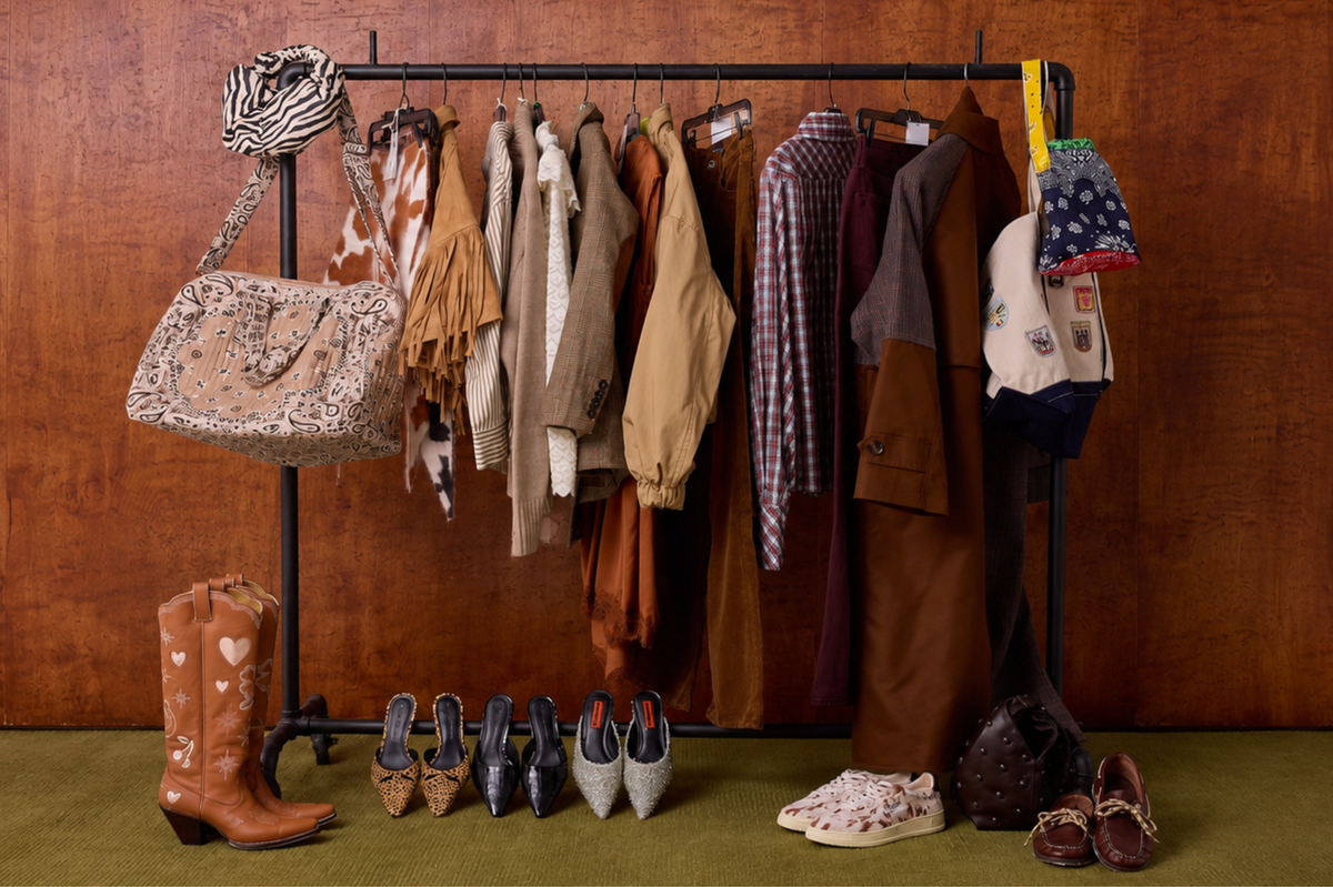A clothing rack filled with multiple garments in earthy tones, styled with a leopard-print shoulder bag, brown leather cowboy boots, pointed flats, and loafers on a green floor against a wood-paneled wall.